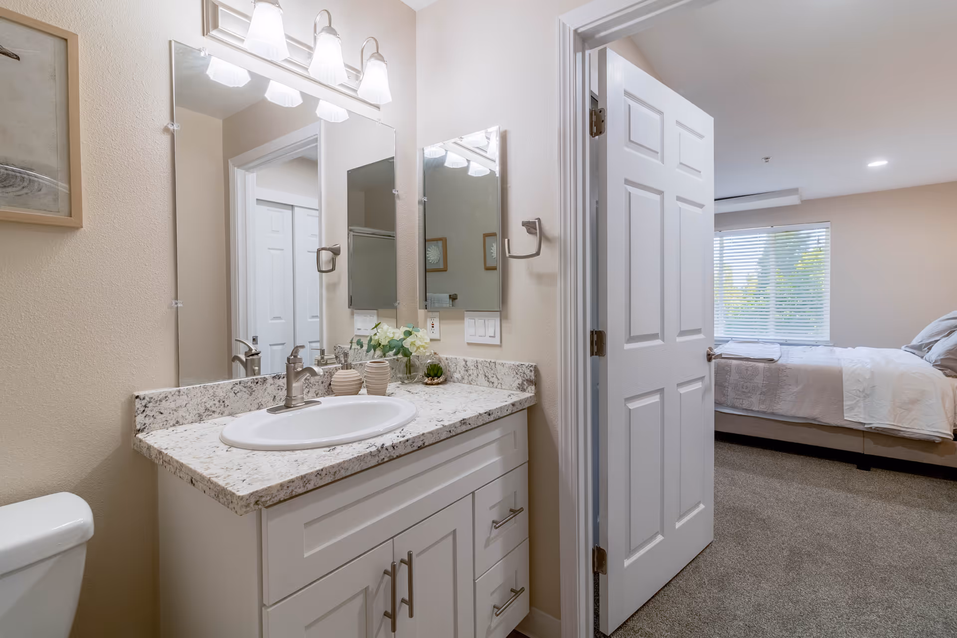 A bathroom vanity with a granite countertop, white sink, and silver faucet. Above the vanity is a large mirror with three light fixtures. To the left, a white toilet is partially visible. The bathroom door is open, revealing a bedroom with a bed, pillows, and a window with blinds.