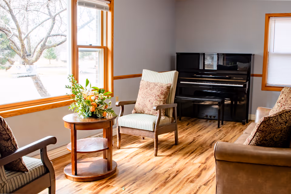 A cozy living room area with wooden flooring, a black upright piano against the wall, a small round wooden table with a floral arrangement, two armchairs with patterned cushions, and a sofa with matching cushions. Large windows with wooden frames allow natural light to fill the room.