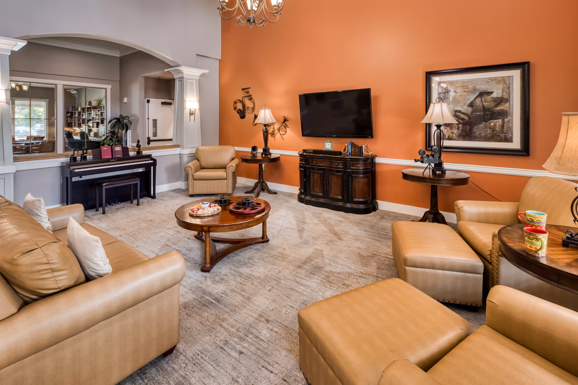 A cozy living room with tan leather sofas and armchairs arranged around a wooden coffee table. The walls are painted orange and gray, with a flat-screen TV mounted on the orange wall above a dark wooden cabinet. There are two side tables with lamps and decorative items, a framed picture of a piano on the wall, and a black piano against the gray wall near a large window.