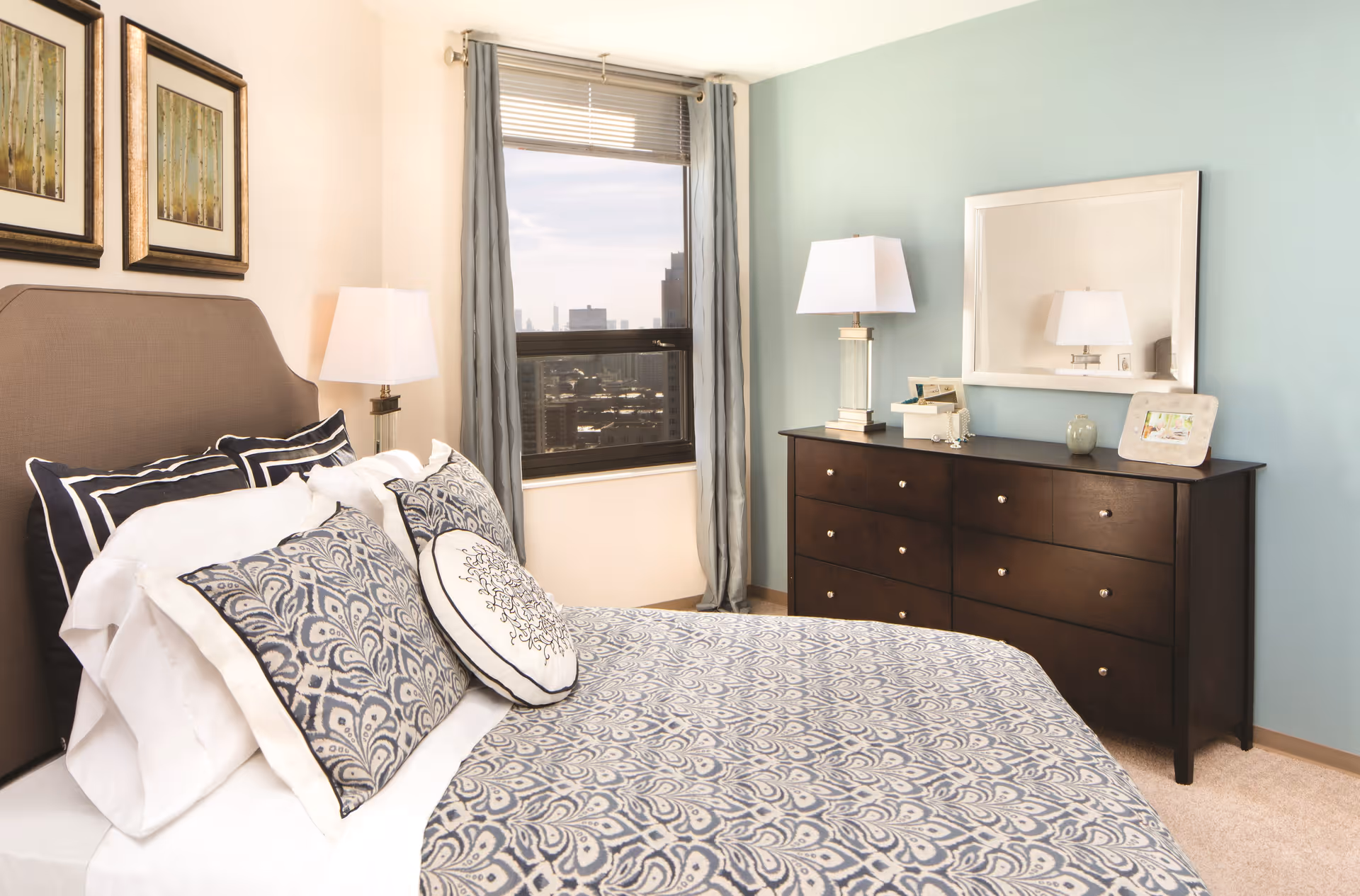 A neatly made bed with patterned blue and white bedding and multiple pillows in a bedroom. The room features a window with gray curtains, two framed pictures on the wall, a dark wooden dresser with a mirror above it, and two table lamps on either side of the bed and dresser.