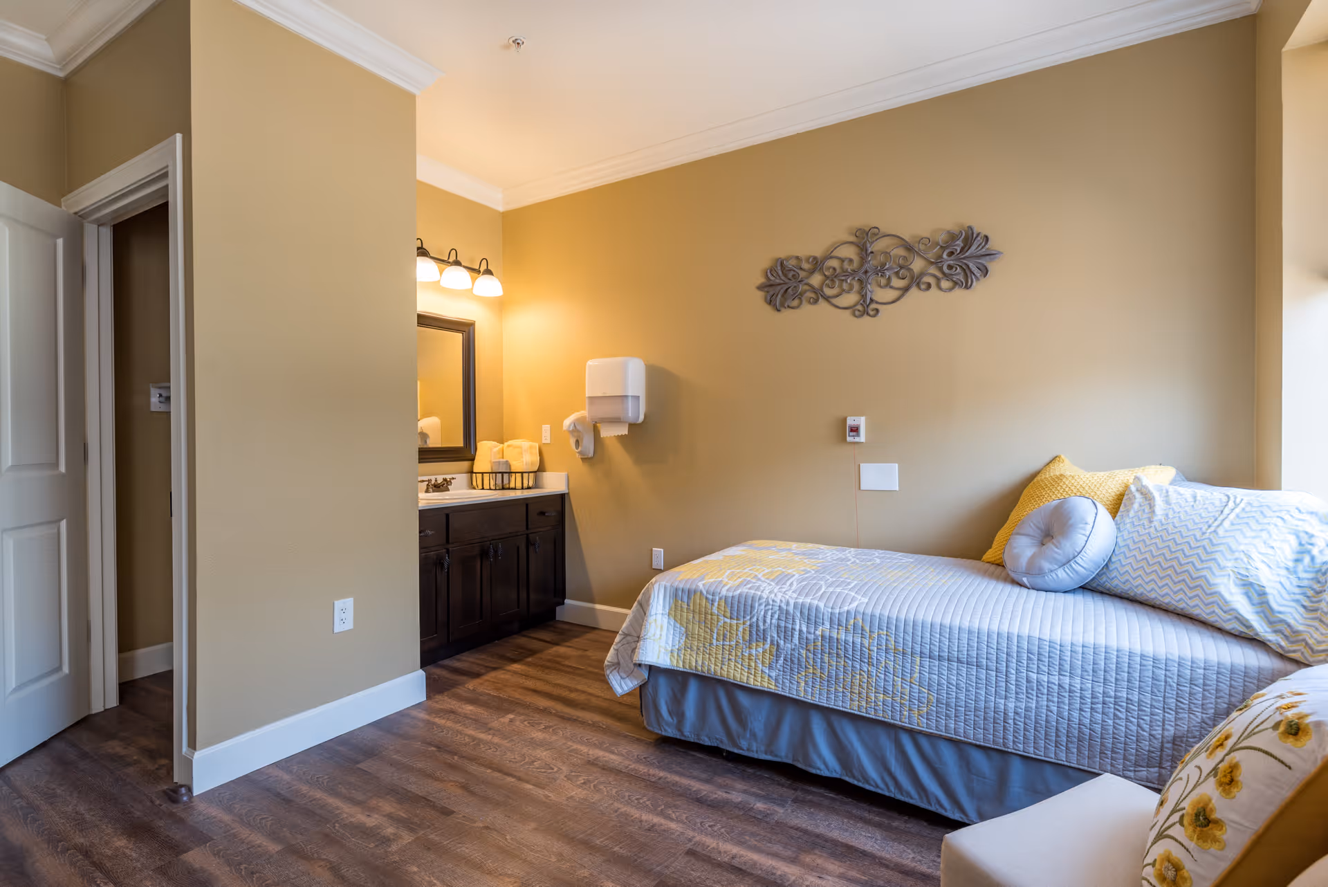 A cozy bedroom with a single bed covered in a light blue and yellow floral quilt, accompanied by several pillows. The room has beige walls, wooden flooring, and a decorative metal wall art above the bed. To the left, there is a vanity area with a mirror, sink, and lighting fixtures, as well as an open door leading to another room.