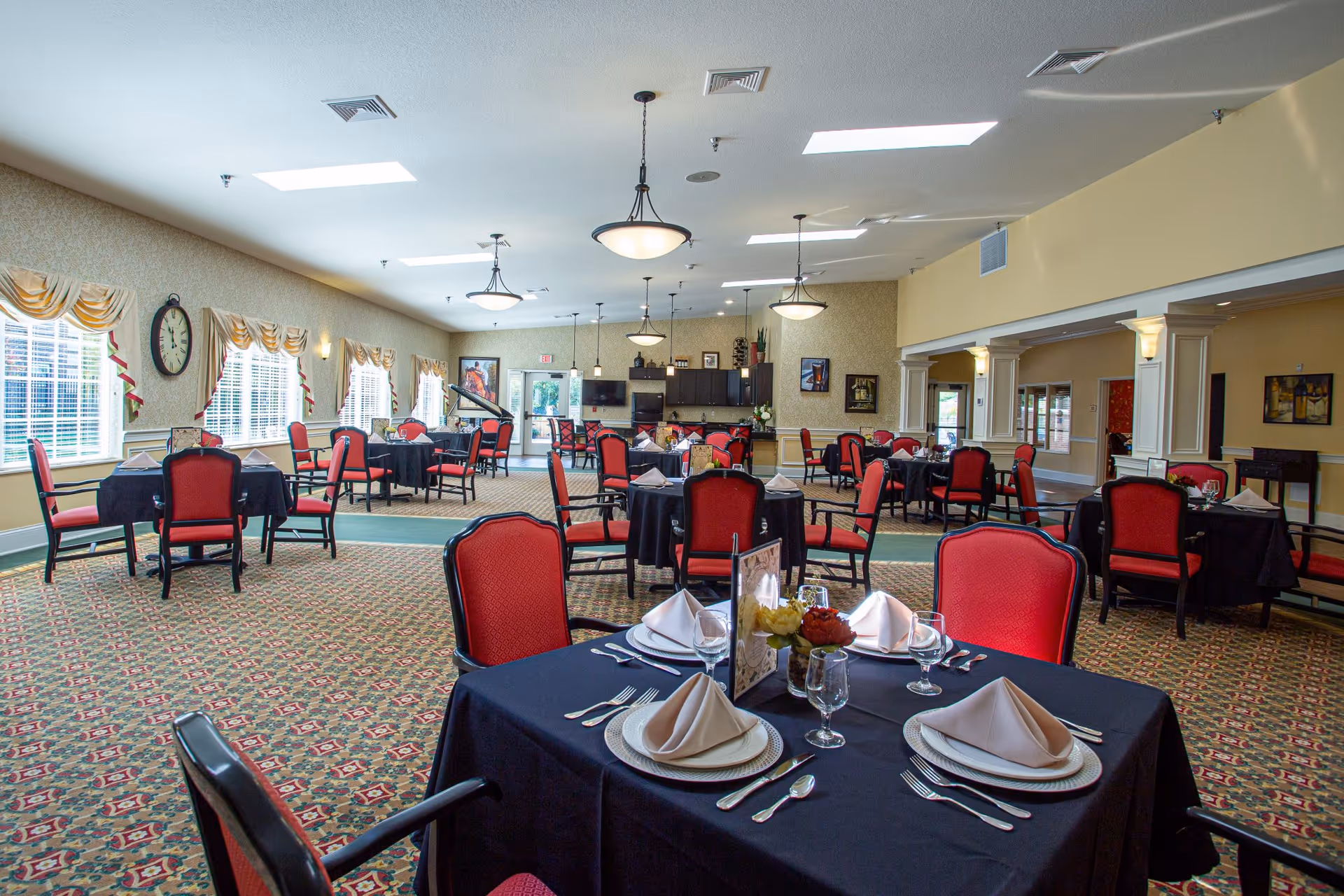 Spacious dining room with multiple set tables covered in black linens, red chairs, and a piano in the background under bright ceiling lights.