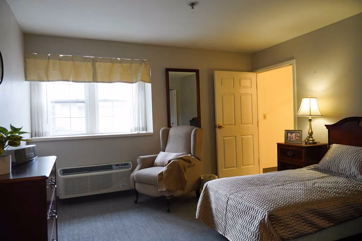 A cozy bedroom in a senior living facility with a bed covered in a textured gray bedspread, a wooden nightstand with a lamp and a framed photo, a beige armchair with a pillow and blanket, a window with sheer curtains and a valance, and a dresser with a plant and a clock radio.