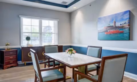 A dining room with a rectangular table surrounded by six chairs with light blue upholstery. The room has a large window with white blinds, a sideboard with decorative items, and a painting of a waterfront scene on the wall. The walls are painted light gray with a blue accent below the chair rail, and the ceiling has recessed lighting.