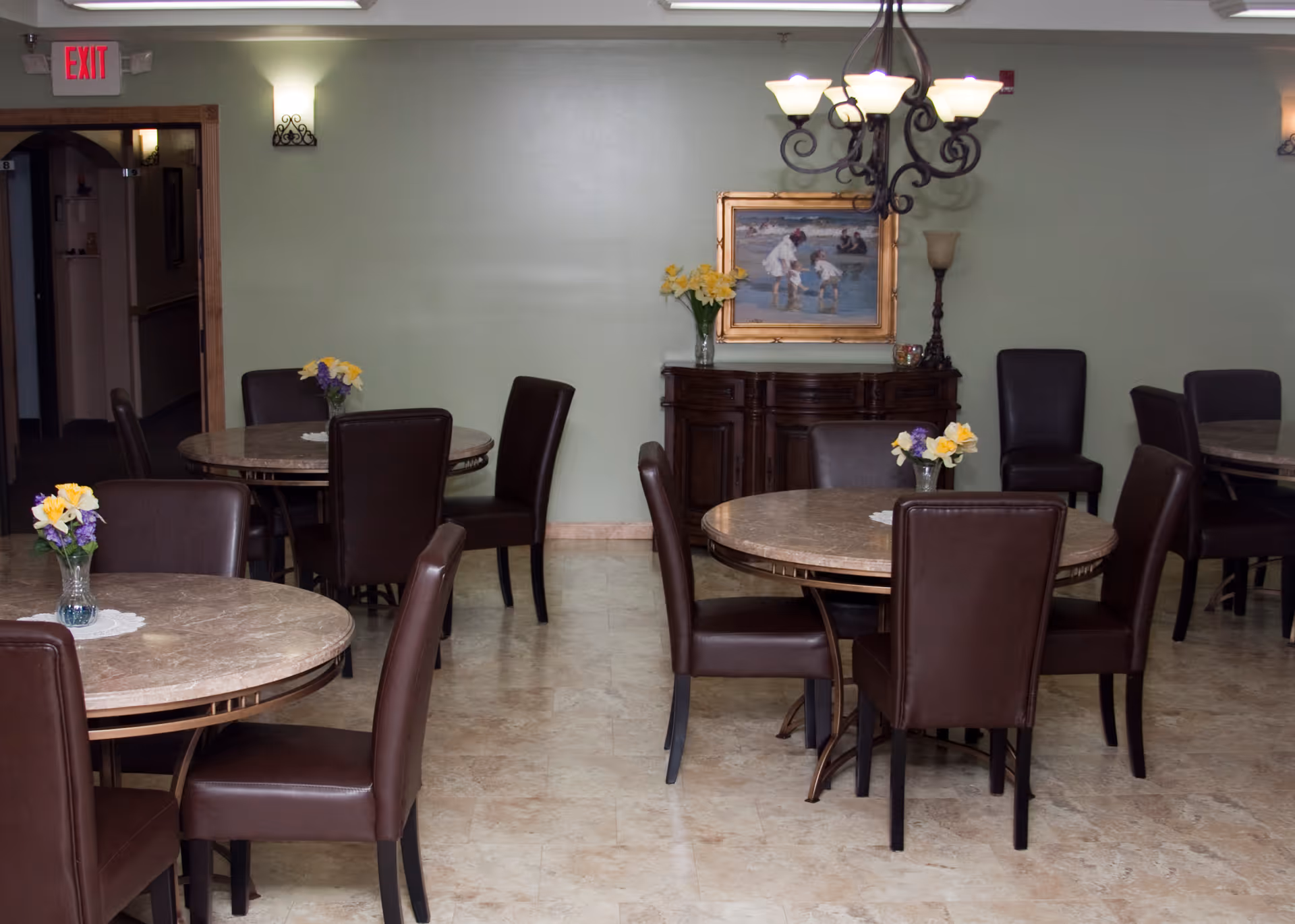 A dining room in a senior living facility with several round tables surrounded by brown leather chairs. Each table has a small vase with yellow and purple flowers. The room has light green walls, a chandelier hanging from the ceiling, a wooden sideboard with a vase of flowers and a framed painting above it, and an exit sign above a doorway.