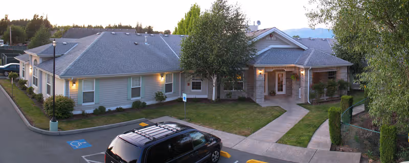 Exterior view of a single-story senior living facility building with a gray roof and beige siding. The entrance is centered with a covered porch and lit wall lamps. There is a parking lot in front with a black vehicle parked in a handicapped parking space. Trees and shrubs surround the building, and a sidewalk leads to the entrance.