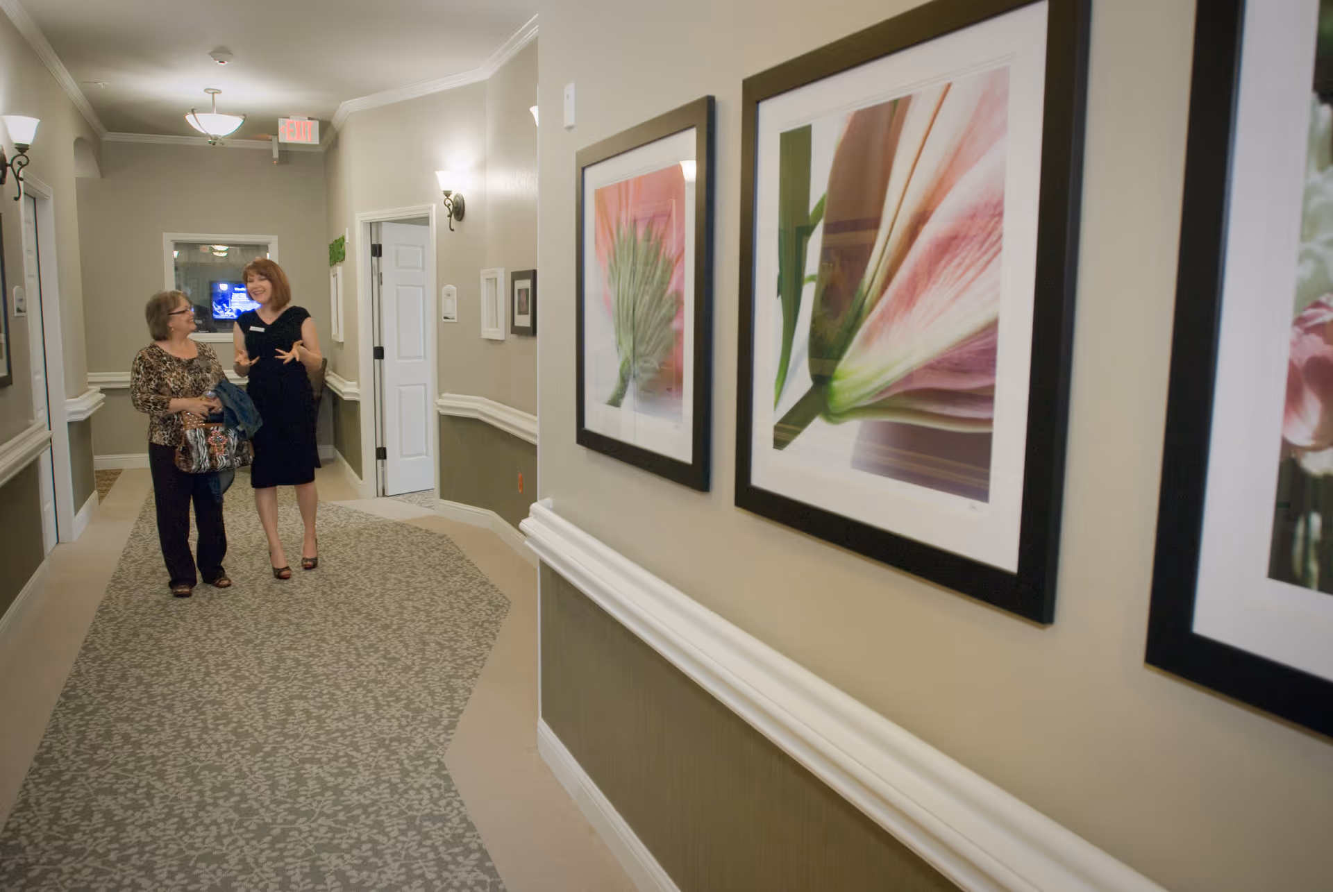 Two women walking and talking in a well-lit hallway decorated with framed floral artwork on the walls and patterned carpet flooring.