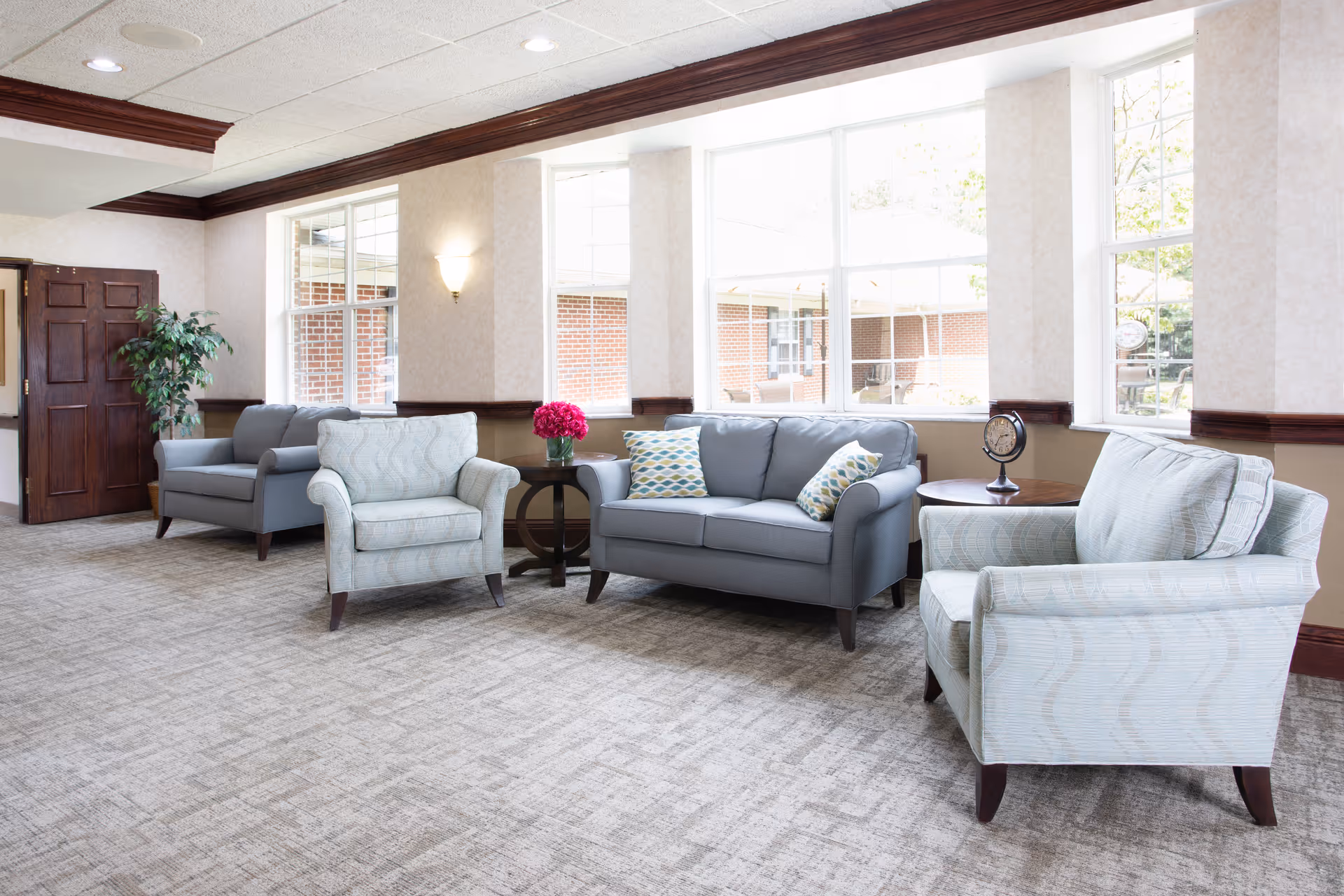 A bright and spacious living room area with large windows letting in natural light. The room features two light blue armchairs, a gray loveseat with patterned cushions, two round wooden side tables, a small decorative globe, and a vase with pink flowers. The walls are light-colored with dark wood trim, and there is a potted plant near a wooden door.