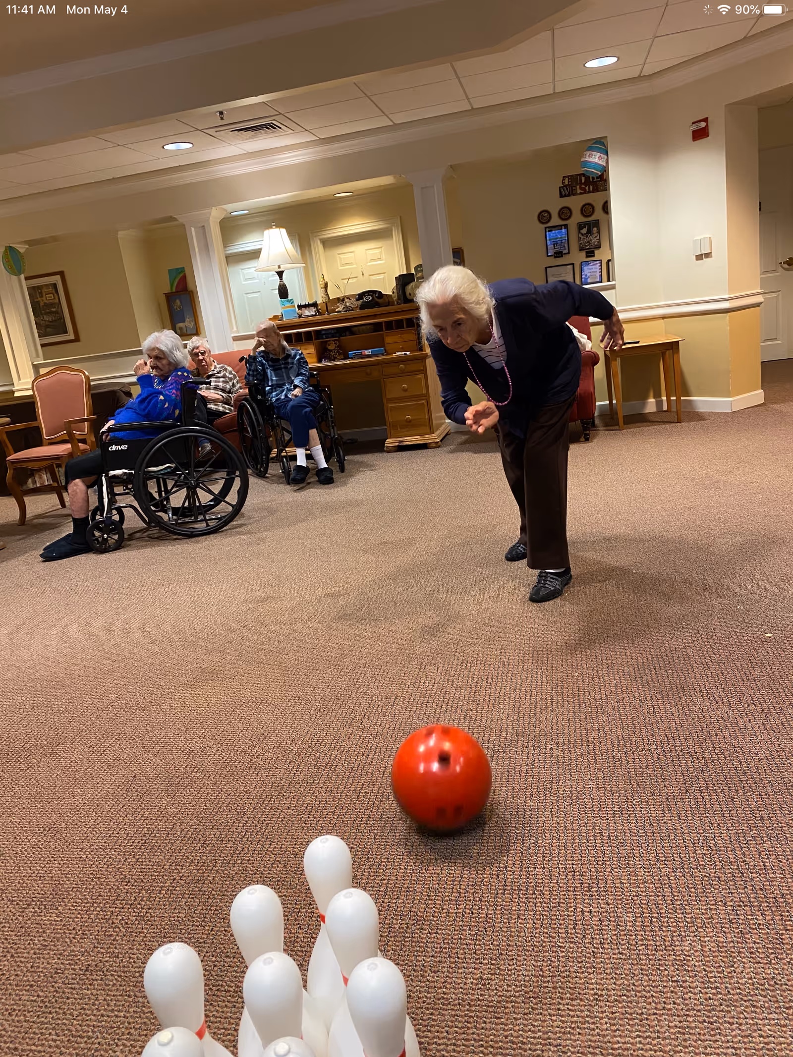 An elderly woman is bowling with a red ball towards white bowling pins set up on a carpeted floor in a common area. In the background, three elderly people, two in wheelchairs and one seated, watch the game. The room has warm lighting, beige walls, and wooden furniture including a desk with a lamp and chairs.
