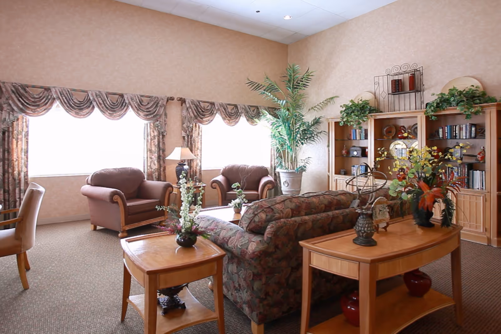 Sunlit living room with a patterned sofa and armchairs, wooden side tables, bookshelves and potted plants.