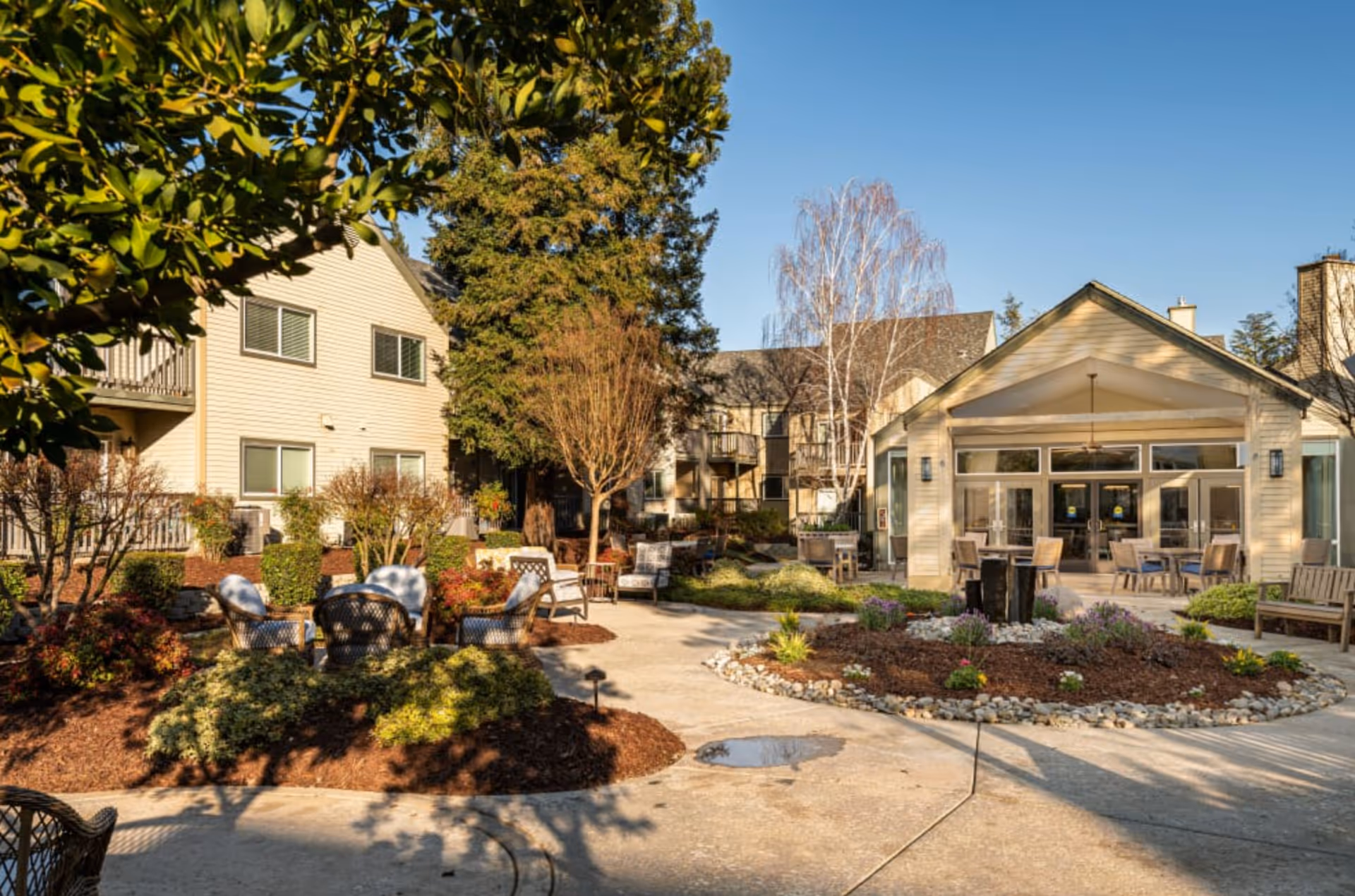 Outdoor courtyard area of Blossom Vale Senior Living featuring a paved walkway, landscaped garden beds with shrubs and flowers, several seating areas with chairs and tables, and a building with large windows and a covered patio in the background under a clear blue sky.