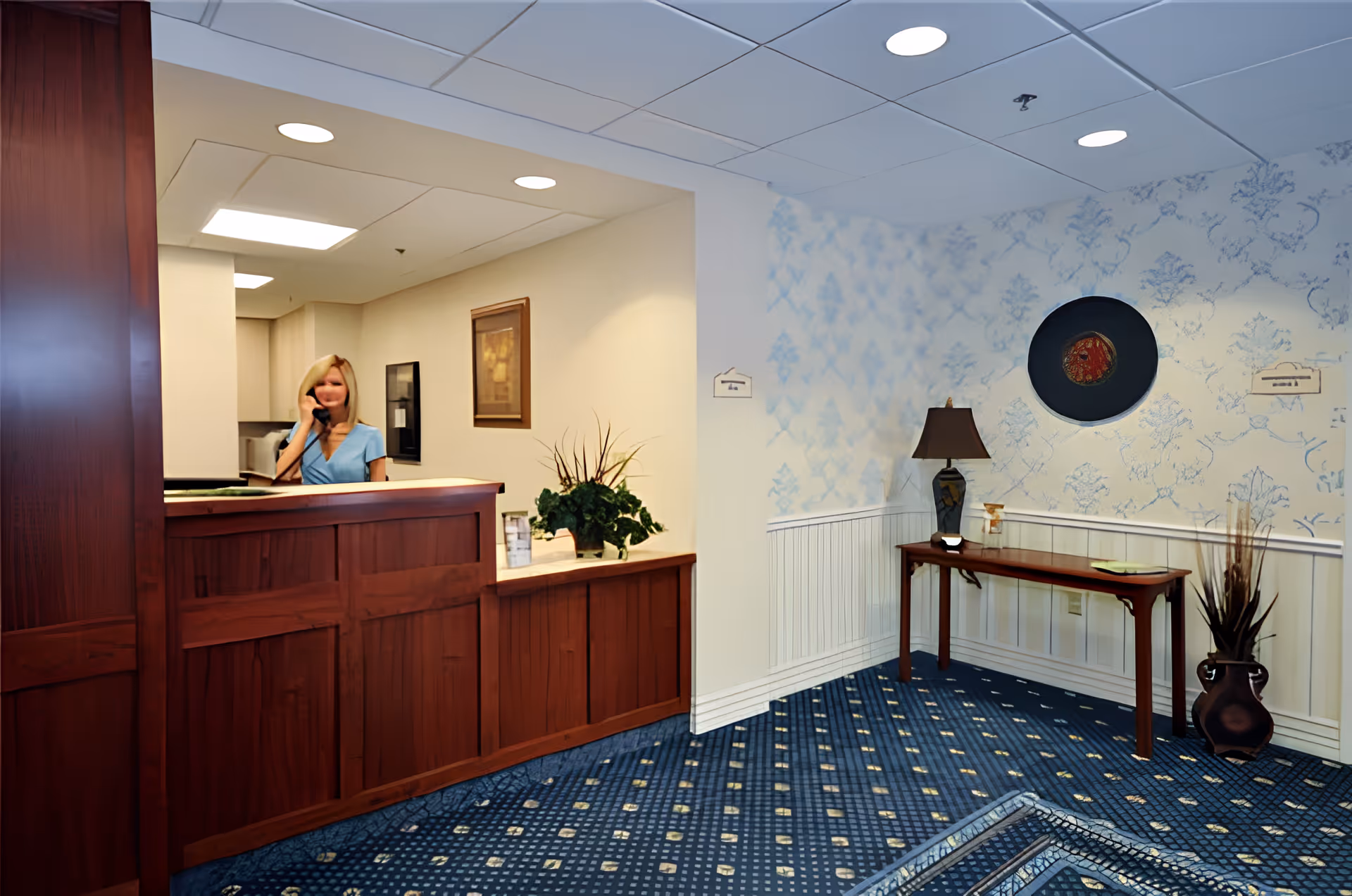 Reception area with a wooden front desk, a woman on the phone, and a decorated seating area with a table and lamp.