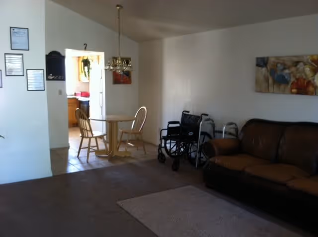 Interior view of a living area in an assisted living facility showing a brown leather couch, two wheelchairs, a small round dining table with two wooden chairs, and a kitchen area visible through an open doorway. The walls are white with some framed pictures and a hanging light fixture above the dining table.