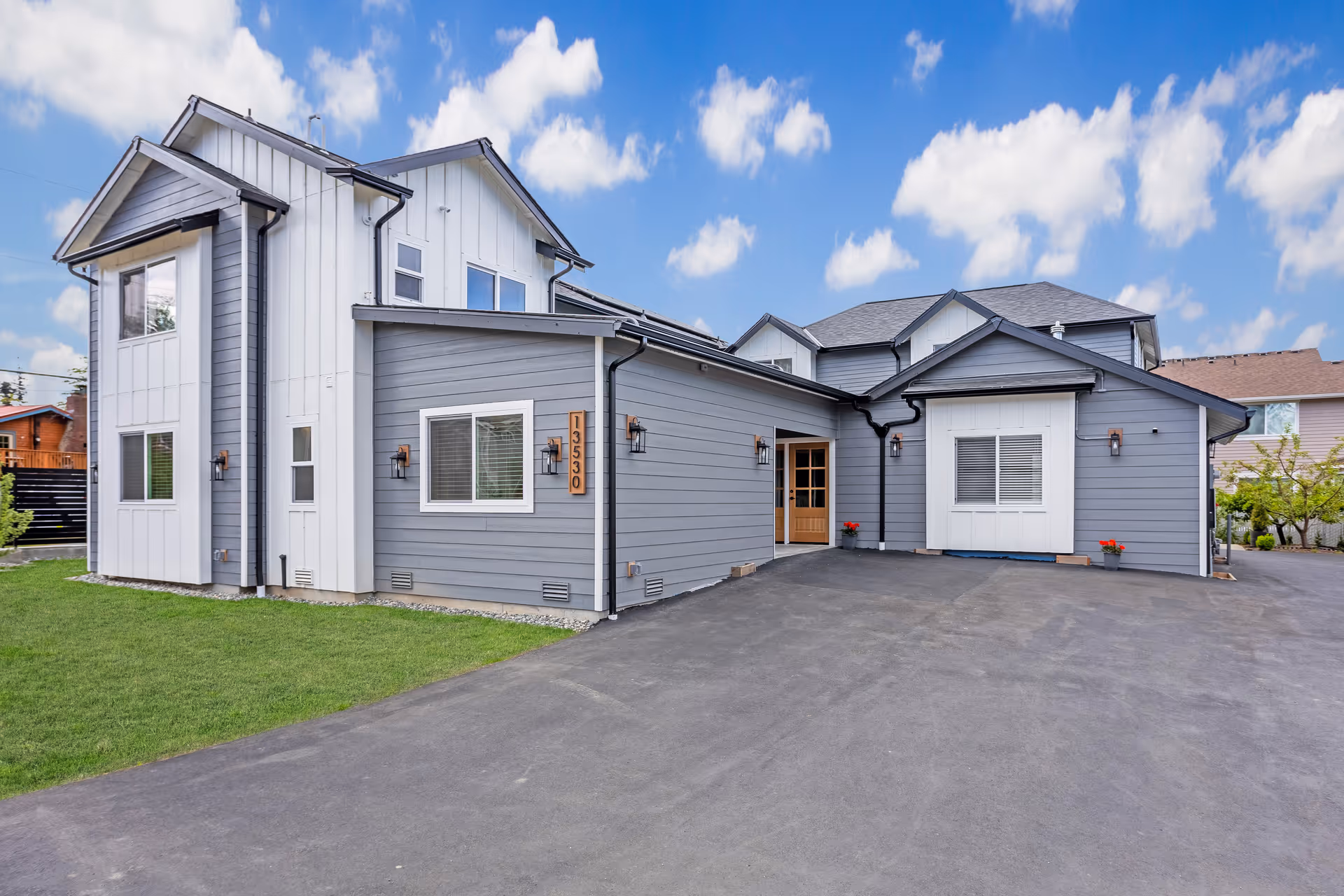 Exterior view of a modern two-story adult family home with gray and white siding, multiple windows, and a paved driveway under a blue sky with scattered clouds.
