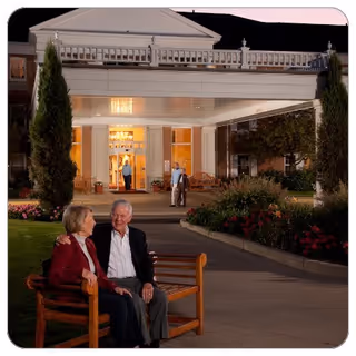 An elderly couple sitting on a wooden bench outside a well-lit building entrance at dusk. The building has a covered driveway with columns and decorative railings above. There are landscaped flower beds and tall shrubs on either side of the walkway. Two other people are visible near the entrance in the background.