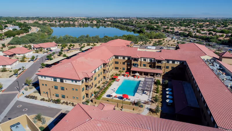 Aerial view of a U-shaped senior living complex with a central swimming pool and courtyard, red-tiled roofs, and a lake in the background.