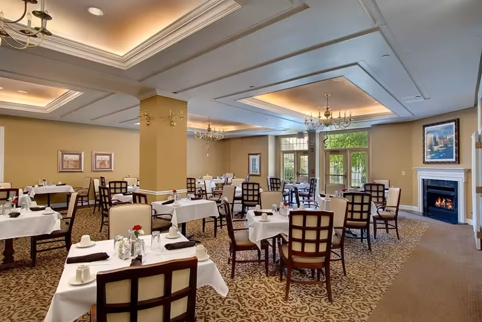 A spacious dining room with multiple tables covered in white tablecloths, each set with cups, napkins, and small flower vases. The room features beige walls, patterned carpet, chandeliers, and a fireplace with a painting above it. Large windows and glass doors allow natural light to enter, showing greenery outside.