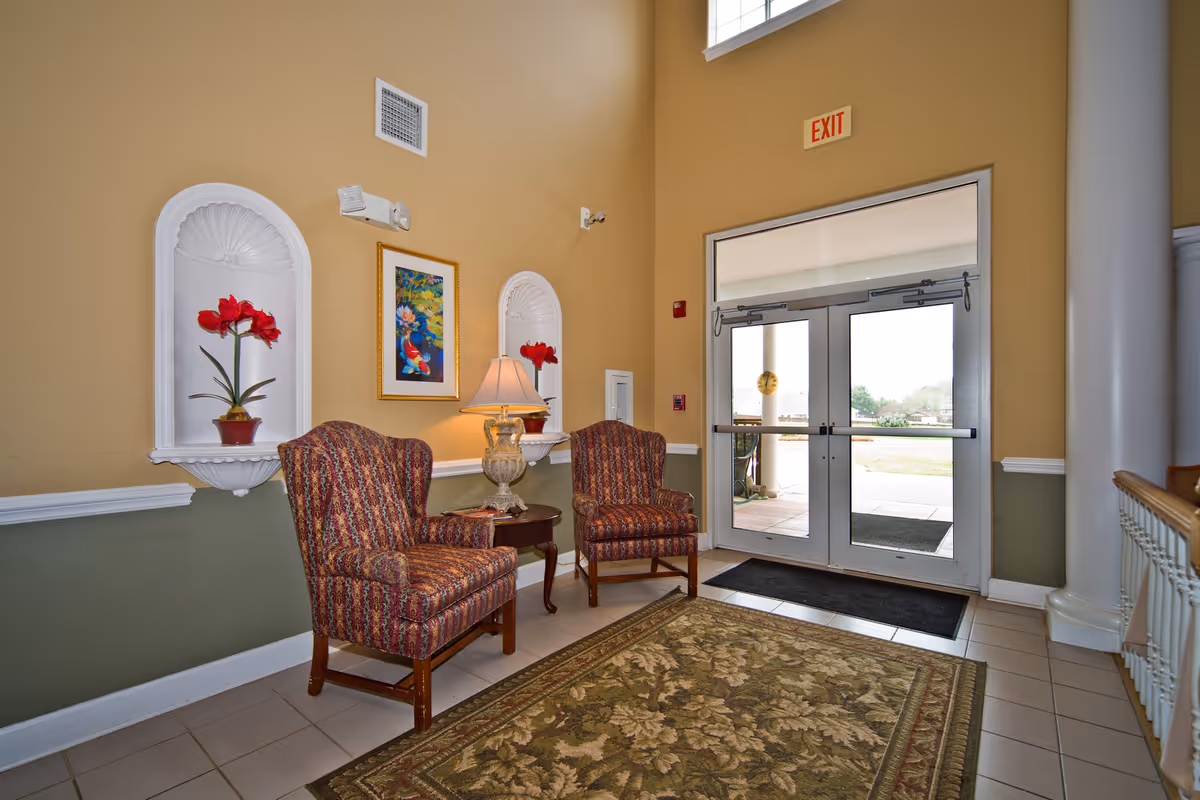 A cozy seating area near the entrance of a facility with two patterned armchairs, a small wooden table with a decorative lamp, two wall niches each holding a red flower pot, a framed colorful painting, and double glass doors leading outside. The walls are painted beige and green, and there is a floral area rug on the tiled floor.