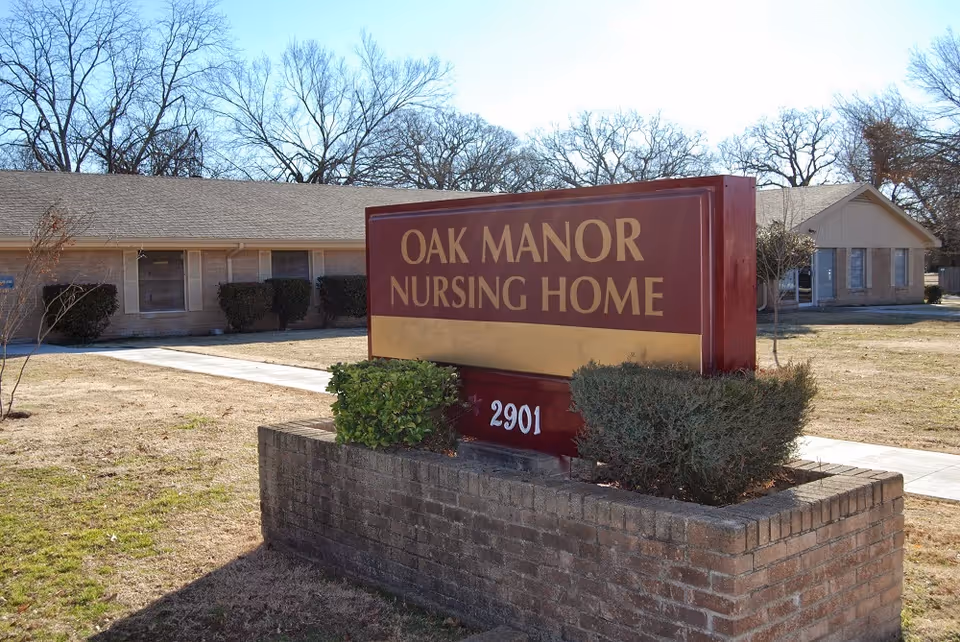 Outdoor view of Oak Manor Nursing Home sign with the building and leafless trees in the background on a clear day.
