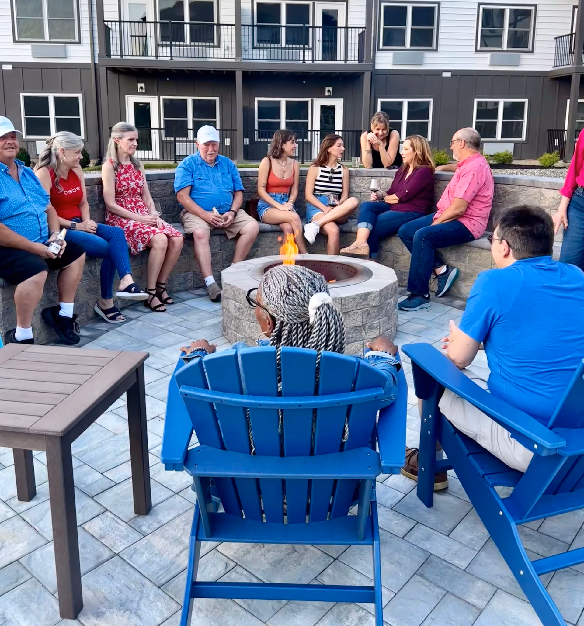 A group of people sitting around a circular outdoor fire pit on a tiled patio. They are engaged in conversation and appear relaxed. The seating includes a stone bench and blue Adirondack chairs. In the background, there is a multi-story building with balconies and windows.