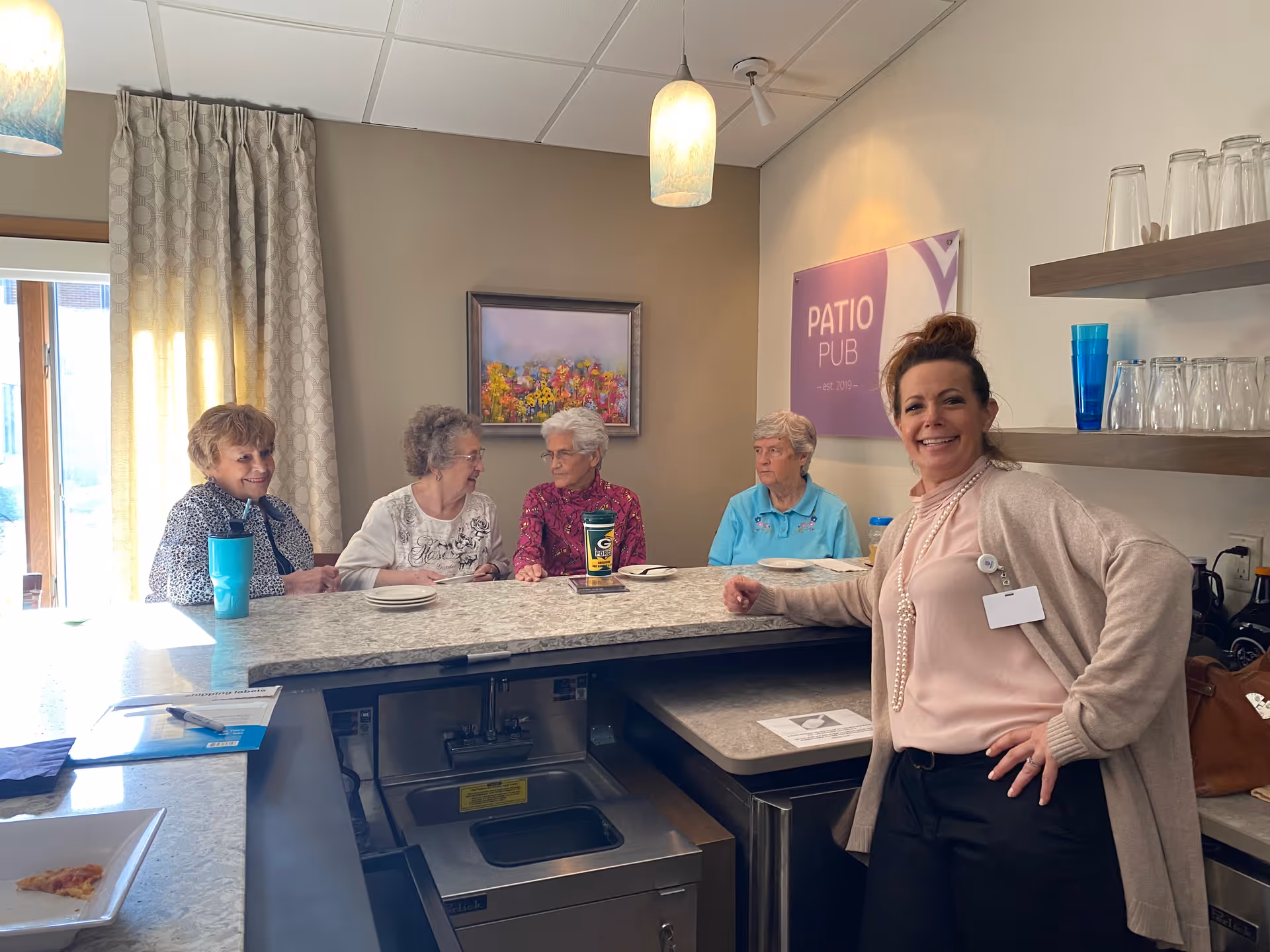 A smiling staff member poses beside four elderly residents seated at a marble counter in a bright common dining area with a 'Patio Pub' sign.