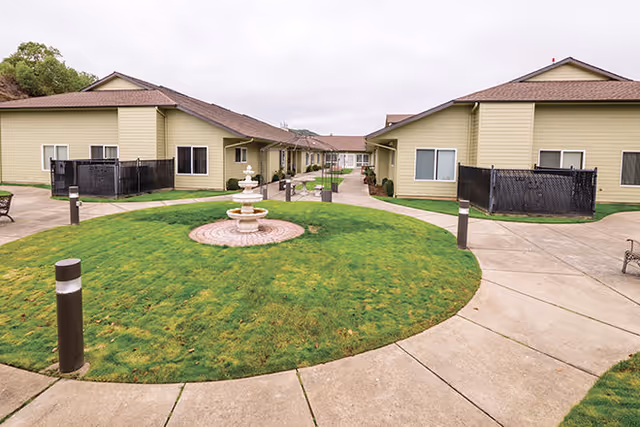 Outdoor courtyard area at Brookdale Roseburg featuring a circular grassy patch with a three-tiered white fountain in the center, surrounded by paved walkways and beige single-story buildings with brown roofs. The sky is overcast.