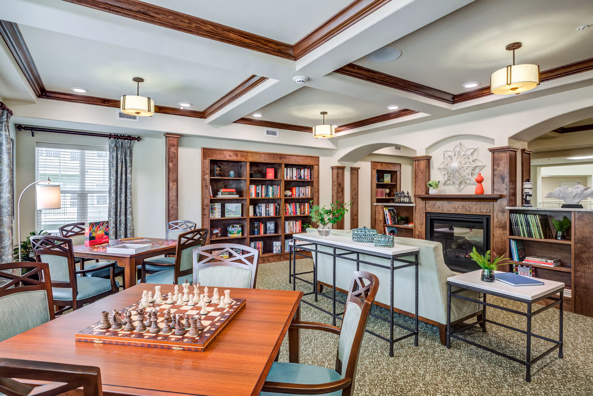 A cozy and well-lit common area in a senior living facility featuring a wooden chessboard on a table in the foreground, several tables and chairs, bookshelves filled with books and games, a fireplace with decorative items on the mantel, and soft lighting from ceiling fixtures. The room has wood trim accents and patterned carpet.
