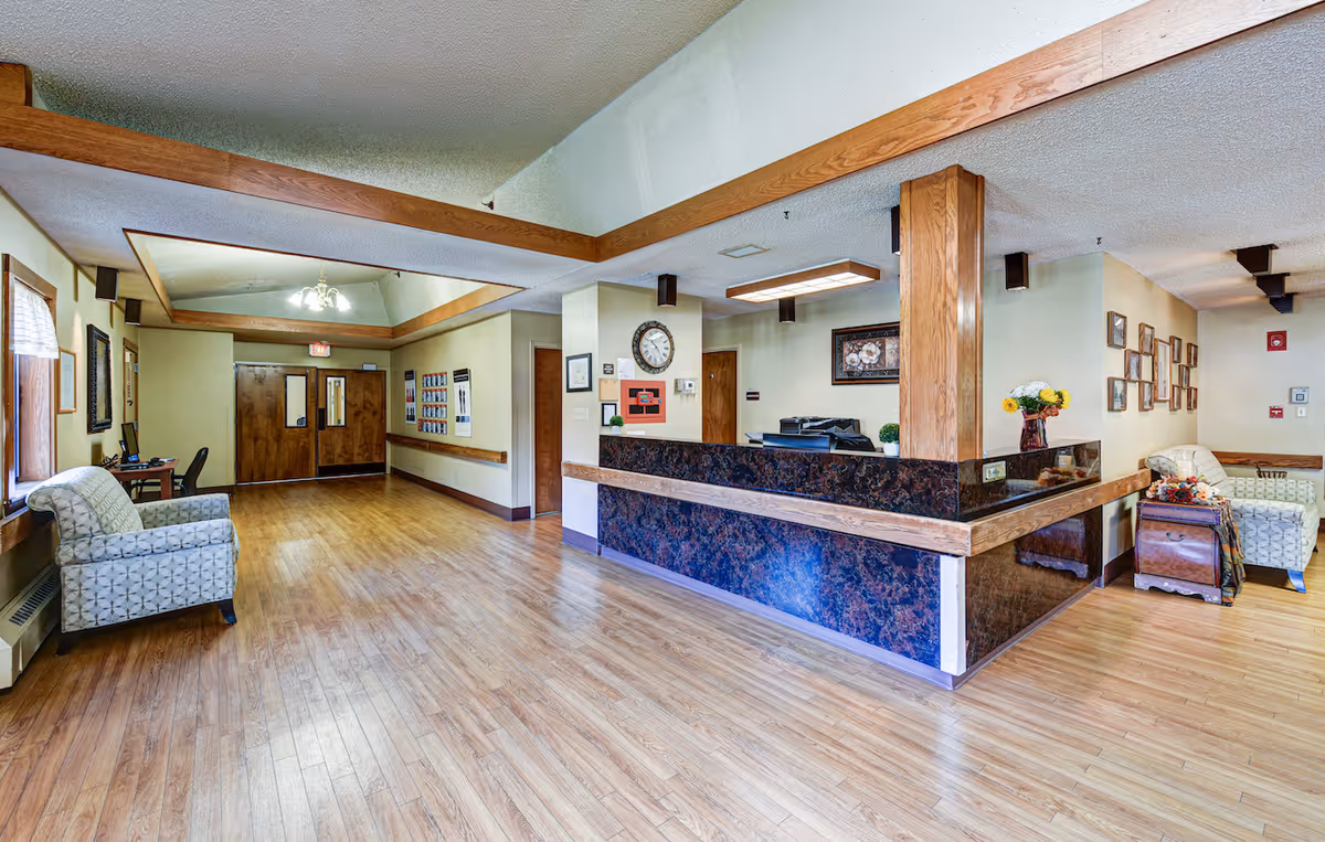 A spacious reception area in a senior living facility with a large wooden and dark marble reception desk. The room has light wood flooring, beige walls, and a ceiling with wooden trim. There are two patterned armchairs on either side of the reception desk, a clock on the wall behind the desk, framed pictures, and a vase with flowers. Double wooden doors are visible at the end of the hallway.