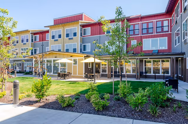 Outdoor courtyard with patio tables, umbrellas, landscaping, and the front of a three-story senior living building.