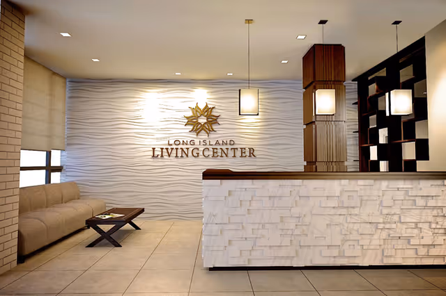 Reception area of Long Island Living Center featuring a modern front desk with textured white panels, a beige couch with a small coffee table, a decorative wall with the facility's name and logo, and hanging pendant lights.