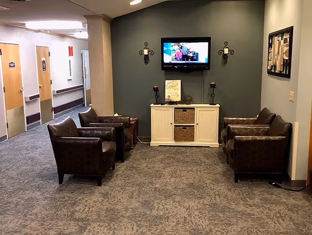 Small communal seating area with four leather armchairs facing a wall-mounted TV above a white storage console in a hallway.