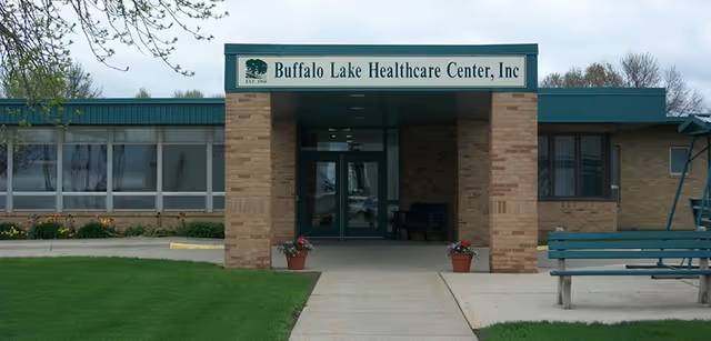Front exterior view of Buffalo Lake Healthcare Center building with a covered entrance supported by brick pillars, a sign above the entrance displaying the facility name, a concrete walkway leading to the door, green grass on either side, and a green bench on the right side.