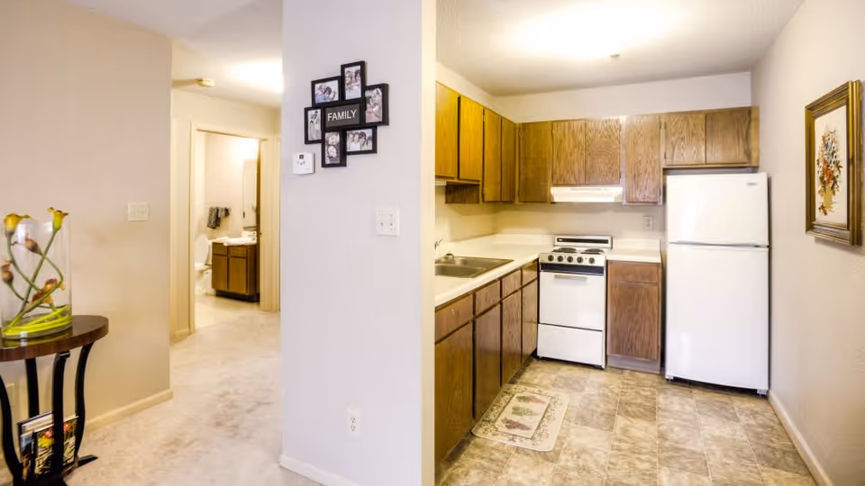 Interior view of a senior living facility apartment showing a kitchen area with wooden cabinets, a white refrigerator, stove, and sink. To the left, there is a small round table with a glass vase containing flowers. A hallway leads to a bathroom with a visible toilet and sink. A photo frame with the word 'FAMILY' is mounted on the wall near the kitchen.