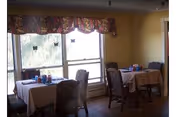 A dining area with two tables covered with beige tablecloths, each surrounded by four upholstered chairs. The tables have condiment bottles and napkin holders. Large windows with floral valances allow natural light to enter the room.