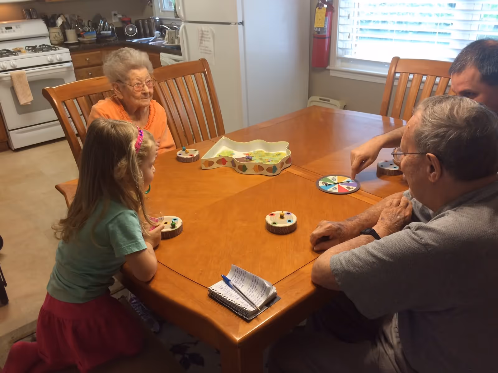 An elderly woman, a young girl, and two elderly men sitting around a wooden dining table playing a board game in a kitchen. The kitchen has a stove, refrigerator, and various kitchen items visible in the background.