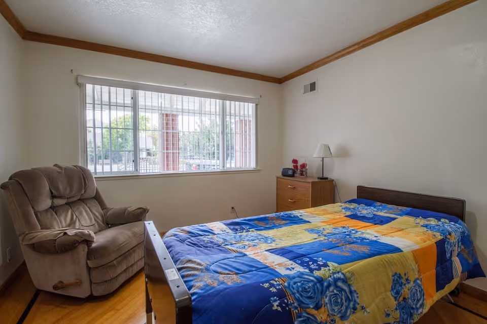 A simple bedroom with a single bed covered in a colorful floral quilt, a wooden nightstand with a lamp and clock radio, a large window with vertical blinds, and a cushioned recliner chair. The room has light-colored walls with wooden trim and a hardwood floor.