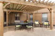 Shaded outdoor patio with a wooden pergola, round table and chairs on a concrete courtyard beside a brick building.