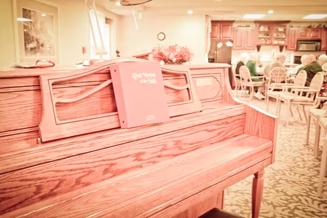 Wooden upright piano with a hymnal on the music rack in a communal dining/activity room where residents sit at tables in the background.