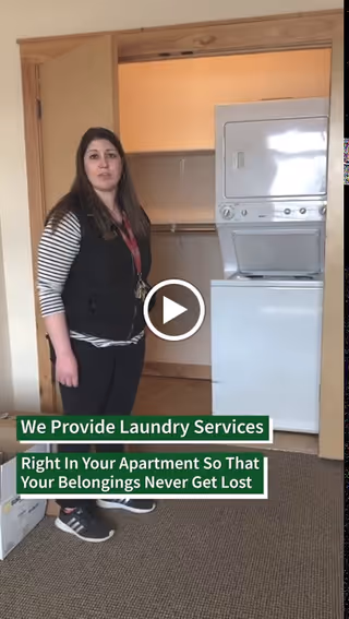 A woman stands next to a stacked washer and dryer unit inside an apartment closet. The closet door is open, revealing the laundry appliances. Text overlay reads: 'We Provide Laundry Services Right In Your Apartment So That Your Belongings Never Get Lost.'