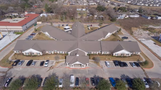 Aerial view of Legend Oaks Healthcare and Rehabilitation Center - Northwest Houston, showing a large single-story building with multiple wings arranged in a Y-shape. The building has a gray roof and beige walls with a covered entrance at the front. Several cars are parked in parking lots in front of and around the building. Surrounding the facility are trees, open land, and residential neighborhoods in the distance.