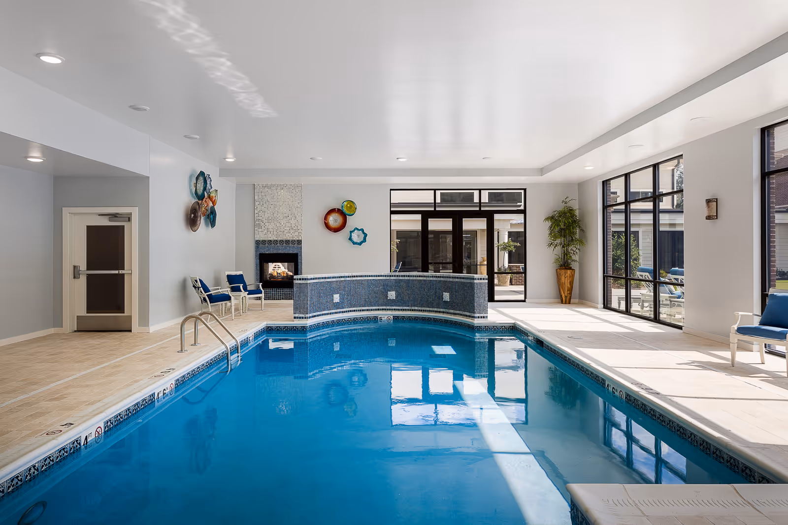 Indoor swimming pool area with blue water, surrounded by beige tiled flooring and white walls. There are blue cushioned chairs along the walls, decorative wall art, a fireplace, large windows letting in natural light, and a potted plant in the corner.