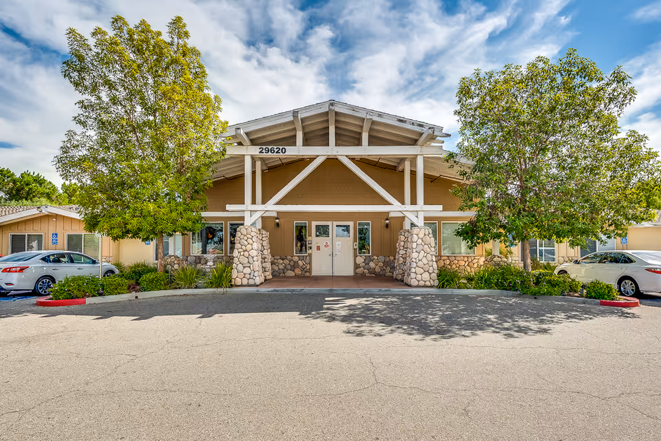 Front entrance of Sunny Rose Senior Living, featuring stone pillars, a peaked roof, and surrounding trees.