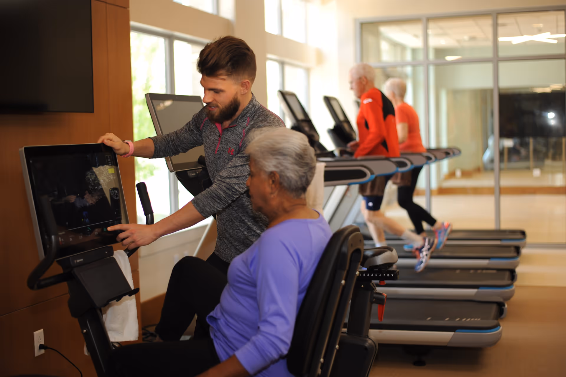 A fitness instructor assists an elderly woman on a stationary exercise bike in a gym. In the background, two elderly individuals are walking on treadmills. The gym has large windows and mirrors on the walls.