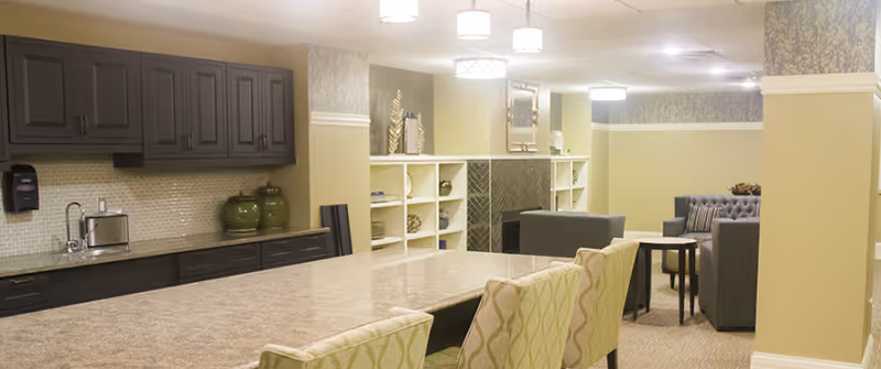 Interior view of a senior living facility common area with a large kitchen island in the foreground, upholstered chairs around the island, dark wood cabinets, a countertop with a toaster and decorative vases, and a seating area with sofas and a coffee table near a fireplace in the background.
