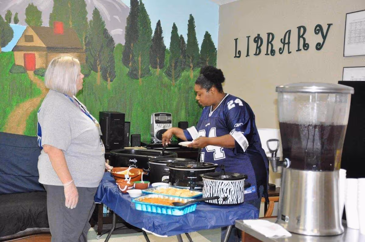 Two women standing at a table covered with a blue tablecloth, serving food from multiple slow cookers and trays in a room with a mural of a house and trees on the wall. The word 'LIBRARY' is displayed on the wall behind them. One woman is wearing a gray shirt and gloves, and the other is wearing a blue sports jersey. A large beverage dispenser with a dark liquid is visible in the foreground.
