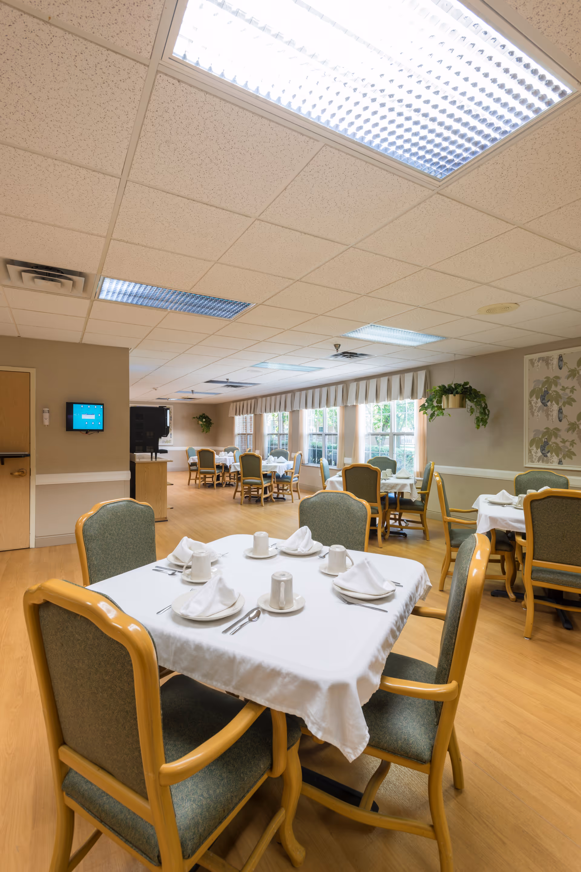 A dining room in a skilled nursing facility with several tables covered in white tablecloths, each set with cups, saucers, napkins, and silverware. The room has wooden flooring, green cushioned chairs with wooden frames, large windows with valances allowing natural light, and some potted plants on the walls.