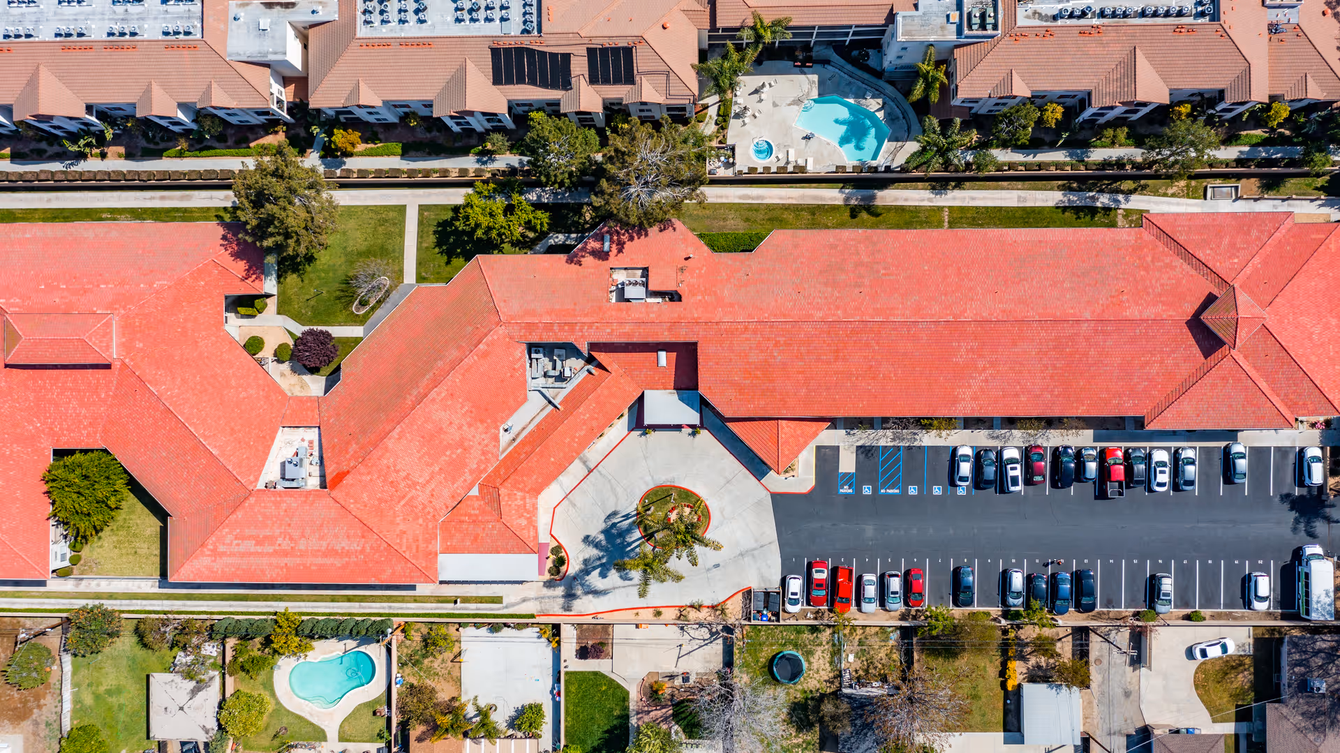 Aerial top-down view of Magnolia Grand showing red-tiled roof buildings, a central driveway entrance, parking lot, pools, and surrounding landscaping.