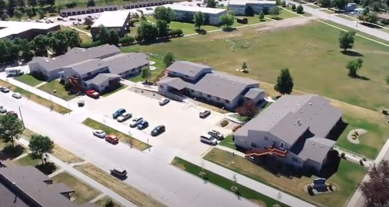Aerial view of a senior living campus with multiple beige buildings, parking lots, and green fields.