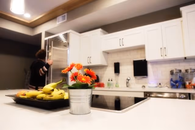 Bright modern kitchen with white cabinets, a central island holding a small potted orange flower and a tray of bananas, and a person opening a stainless refrigerator in the background.