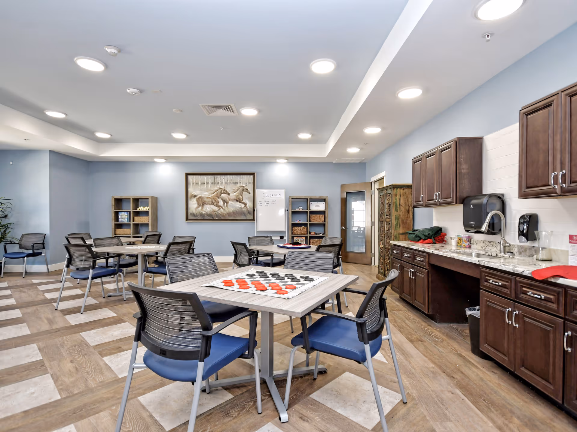 A spacious activity room in a senior living facility with several tables and chairs arranged for group activities. One table has a large checkers game set up. The room features light blue walls, wooden cabinets with a countertop and sink on the right, and shelves with baskets and games at the back. A framed picture of running horses and a whiteboard are mounted on the back wall. The floor has a patterned wood and tile design, and the ceiling has multiple recessed lights.